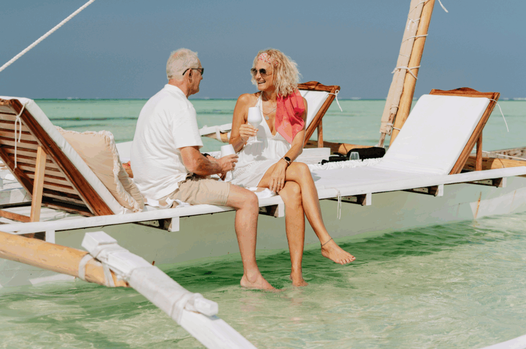 An older man and a woman sit on a wooden boat with lounge chairs, relaxing in clear, shallow turquoise water under a bright sky. Both are smiling and wearing light, summery clothing.