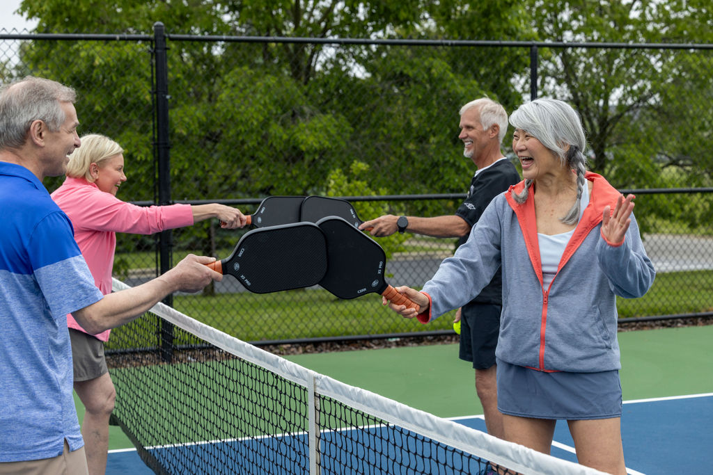 Four older adults standing on opposite sides of a pickleball court, smiling and tapping their paddles over the net in a friendly gesture, with trees and a fence in the background.