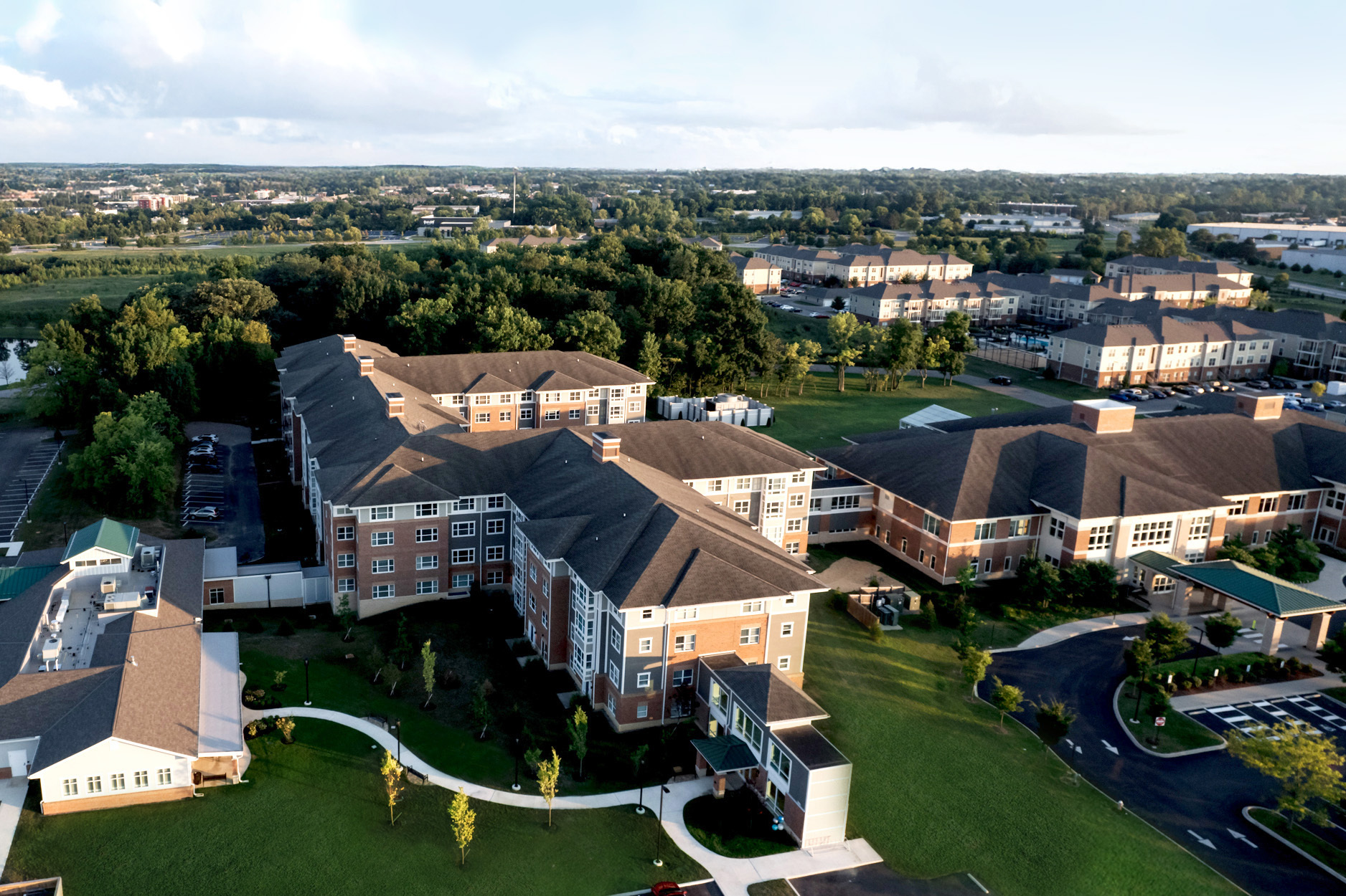 Aerial view of a large residential or institutional complex with multiple connected brick buildings surrounded by green lawns, trees, and pathways under a partly cloudy sky.