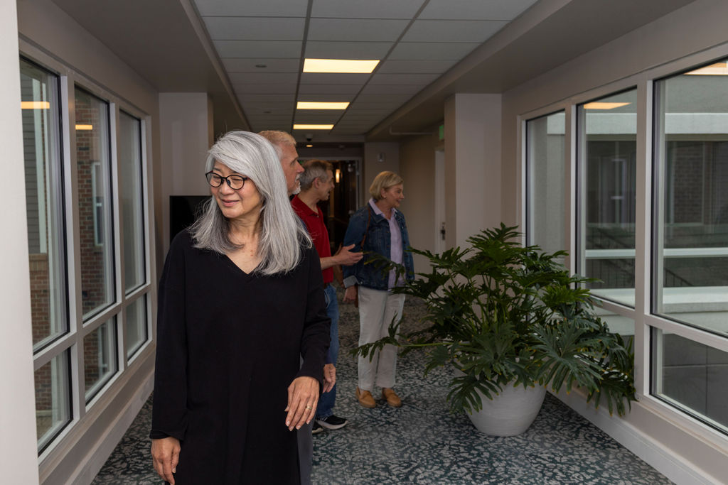 A woman with long gray hair and glasses stands in a hallway with windows, reflecting on small goals as three people in the background look outside. A large potted plant sits on the right.