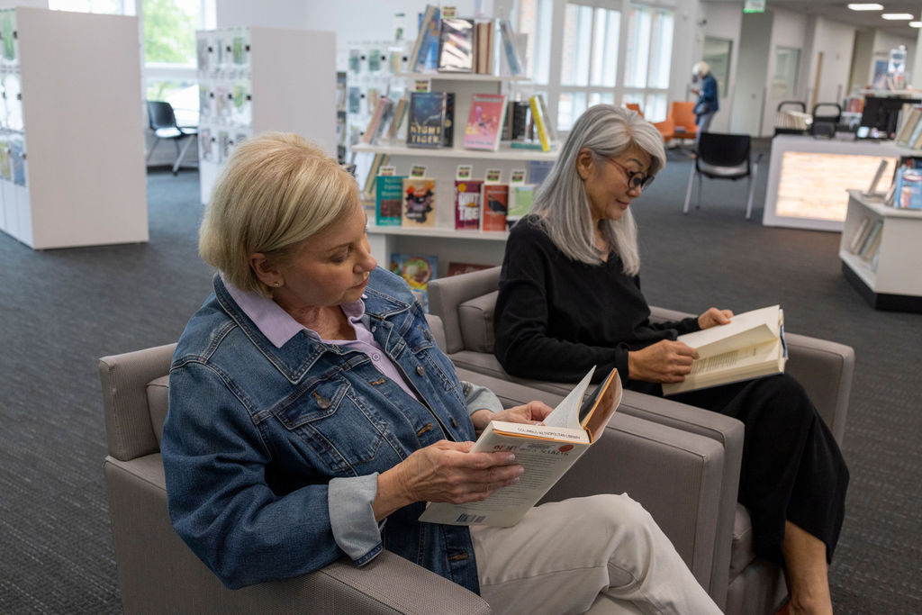 Two women sit in armchairs at a library, each reading a book. Bookshelves and large windows are visible in the background, as they enjoy the calm setting—perfect for reflecting on small goals while immersed in their reading.