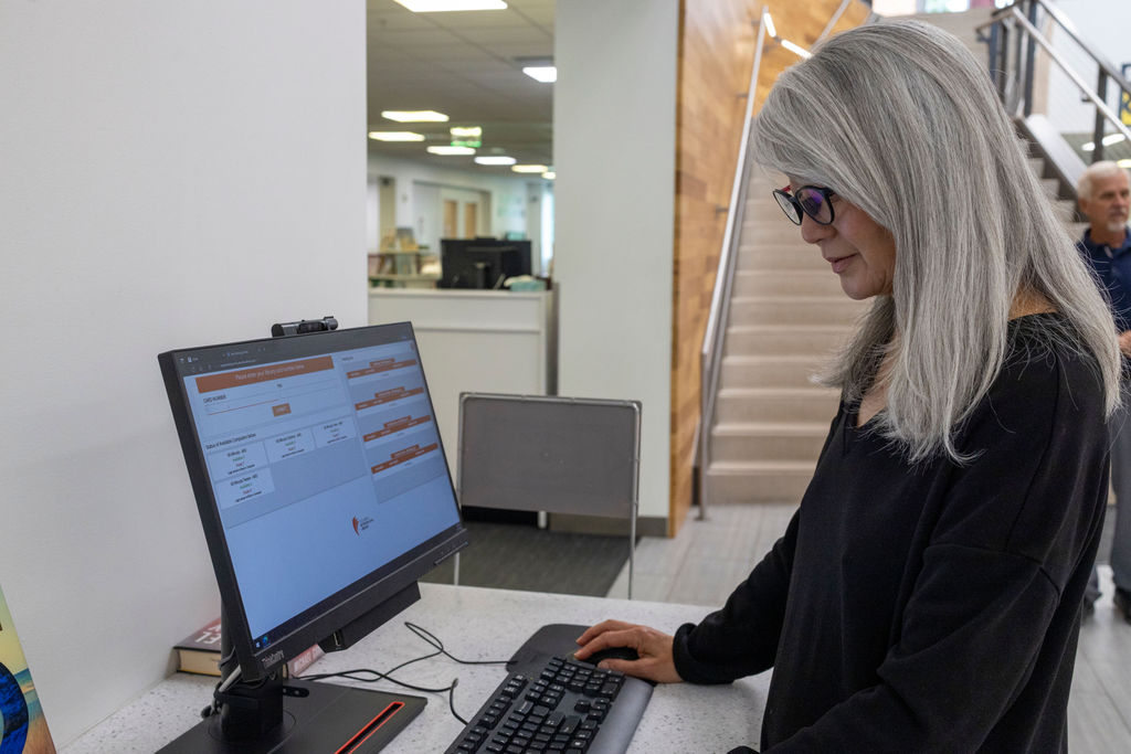 A woman with long gray hair and glasses sets small goals as she uses a desktop computer at a library counter. Stairs and bookshelves line the background, while another person walks by.