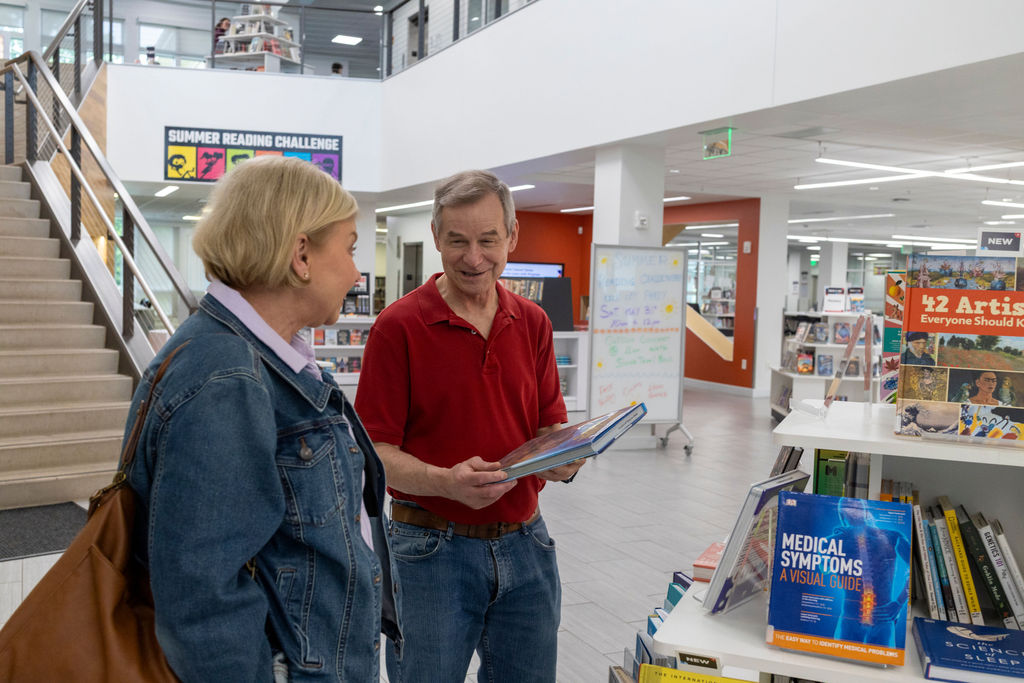Two adults stand in a library near a bookshelf, talking and smiling. One is holding an open book. Books and signs, including one for a summer reading challenge that encourages small goals, are visible in the background.
