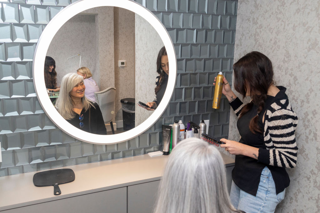 A hairstylist holding a can of hairspray styles a woman with long gray hair, who is seated and looking at her reflection in a round, illuminated mirror on a textured wall. Products are arranged on the counter.