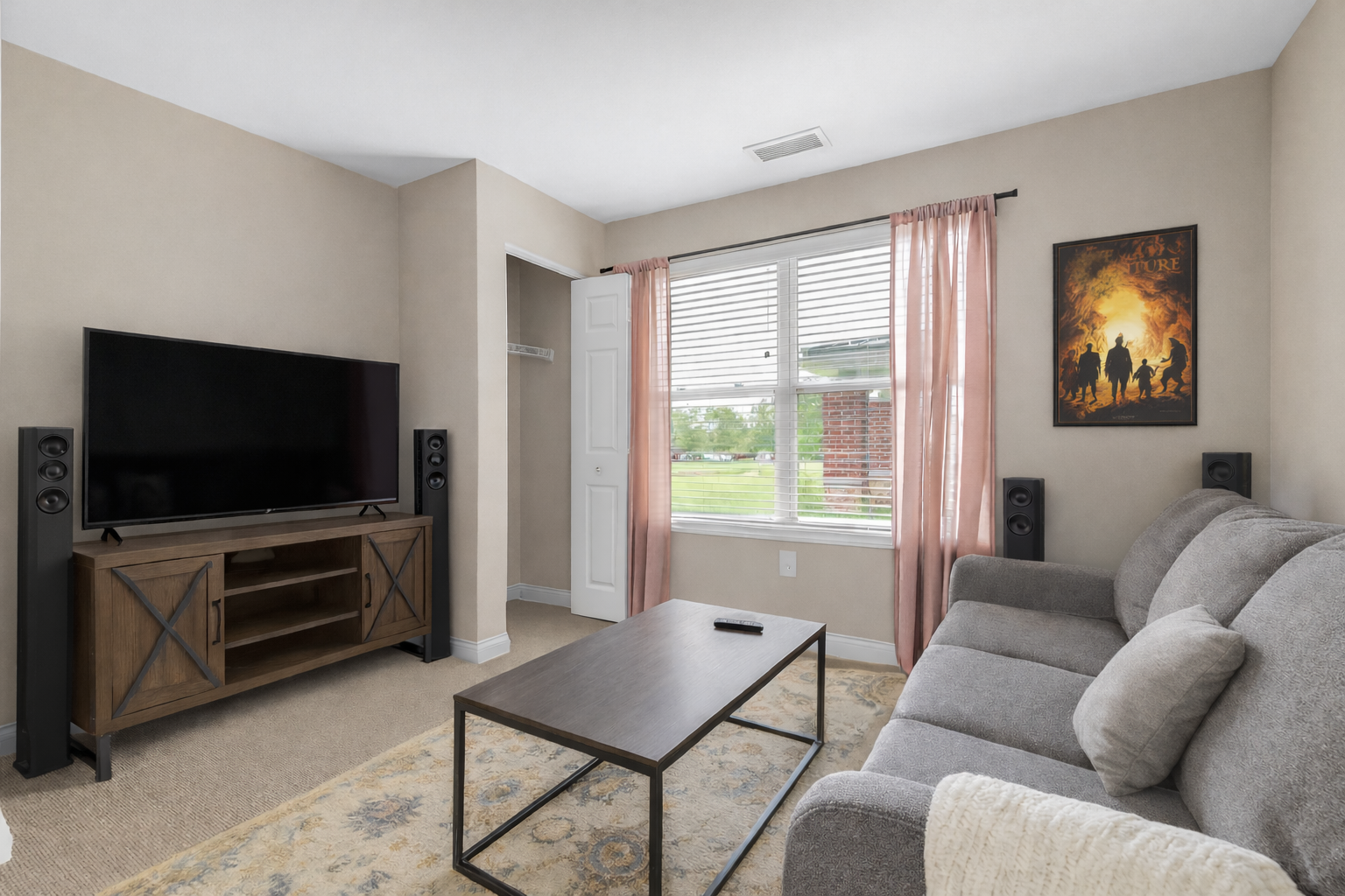 A cozy living room in our spacious living Apartments features a gray sofa, wooden TV stand with flat-screen TV, coffee table, beige walls, a window with pink curtains, and a movie poster on the wall.
