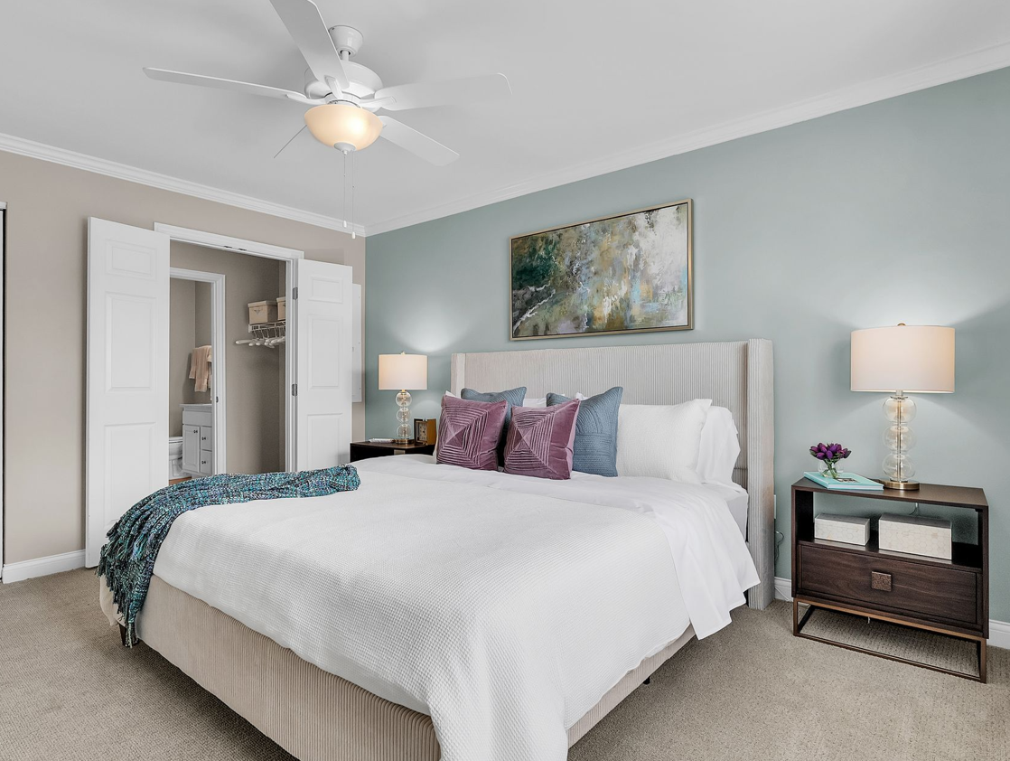 A modern bedroom with a large bed, white bedding, purple and blue accent pillows, and a teal throw awaits in our two-bedroom residences—enjoy more space and style, with a ceiling fan, lamps, art above the headboard, and open access to closet and bathroom.