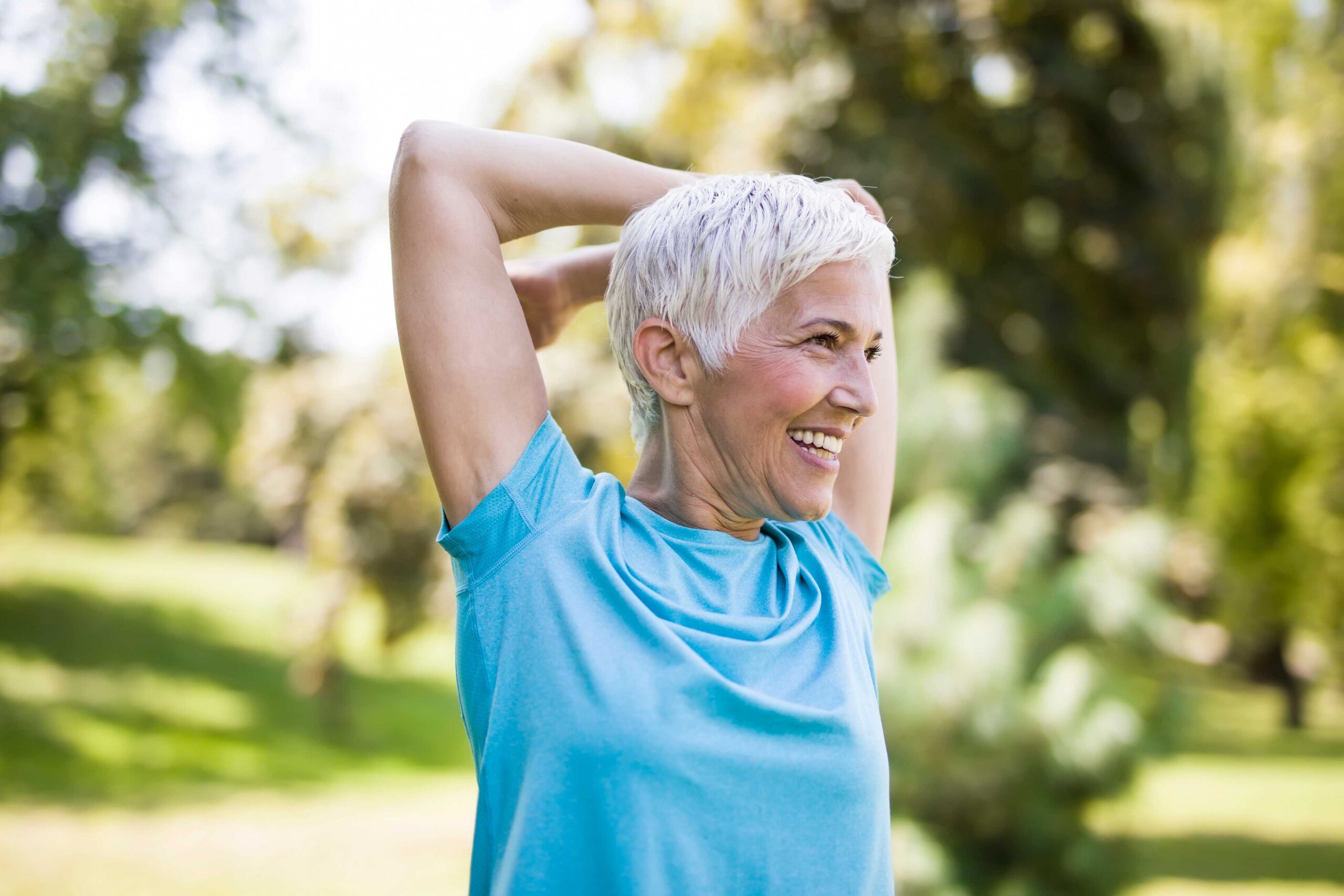 Smiling older woman with short gray hair stretches her arms overhead while standing outdoors in a sunny, green park. She is wearing a light blue shirt and appears relaxed and happy, enjoying the benefits of active partnerships in wellness.