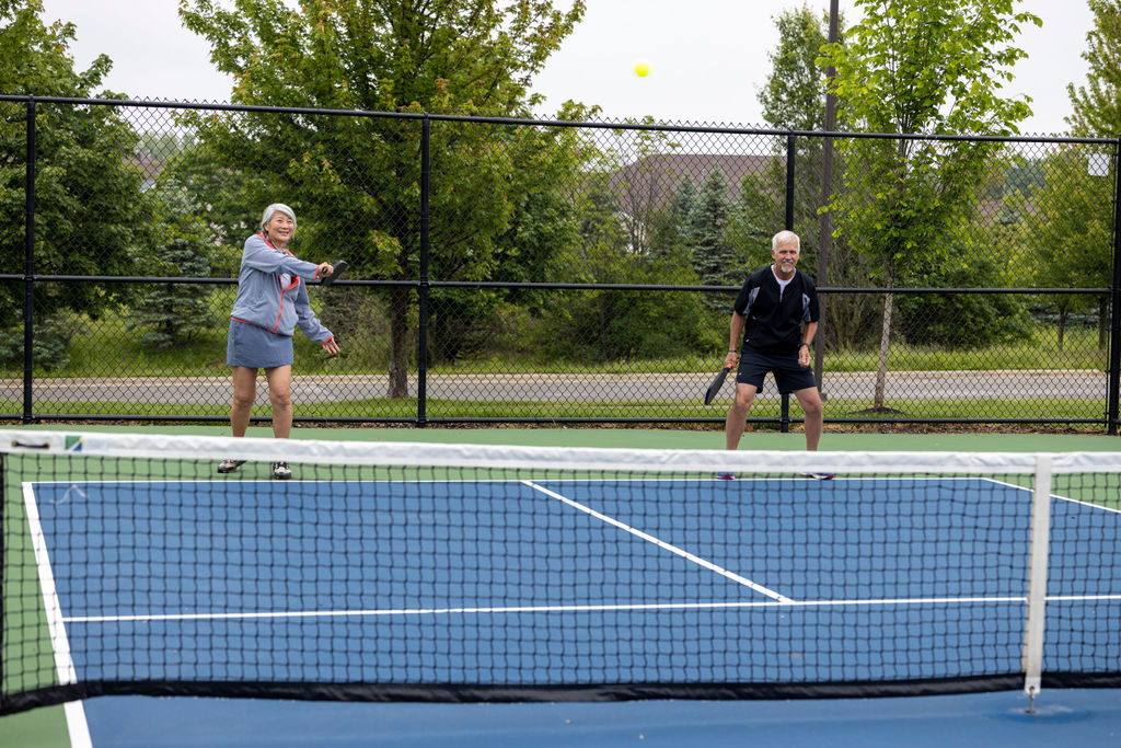 Two older adults enjoy a lively game of pickleball on an outdoor court at Verena at Hilliard. The woman swings her paddle to hit the ball, while the man stands ready behind her. Trees and a fence line the background.
