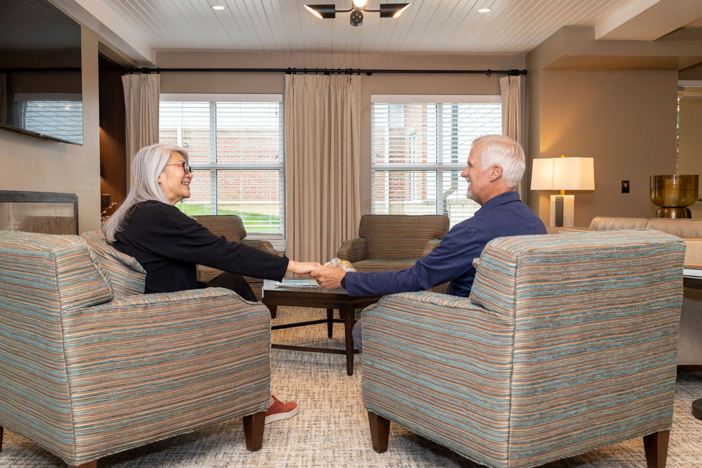 An older woman and man sit in striped armchairs facing each other, holding hands and smiling in a bright, modern living room with large windows—reminiscent of a quiet corner at the Columbus Library—accented by neutral decor.