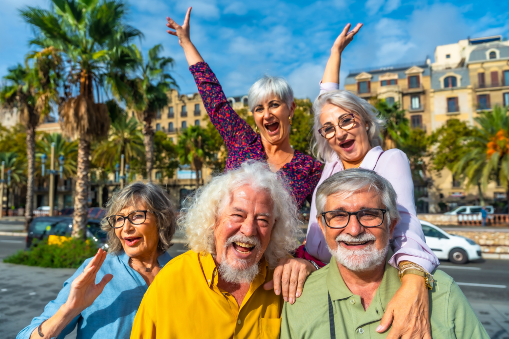 Five joyful older adults pose together outdoors on a sunny day, with palm trees and historic buildings in the background. Three women stand behind two men, all smiling and appearing cheerful and vibrant.
