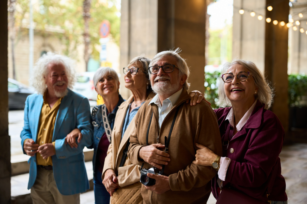 A group of five smiling older adults stands outdoors under a covered walkway, looking up. One person holds a camera, and they all appear happy and engaged, dressed in colorful, stylish clothing.