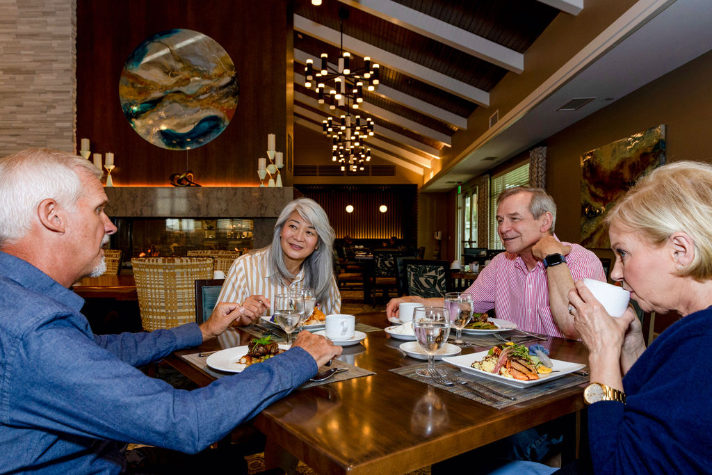 Four older adults sit at a restaurant table enjoying a meal together at Verena at Hilliard. Three are engaged in conversation, while one woman sips from a white cup. The setting is modern and warmly lit with elegant decor.