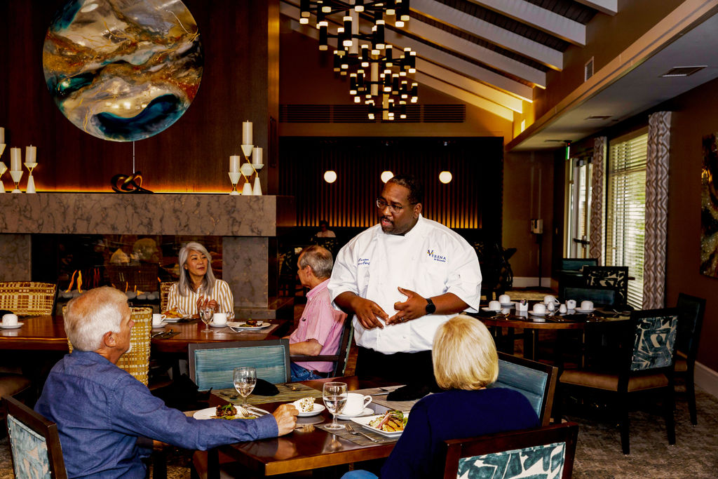 A chef stands and talks to three seated diners at Verena at Hilliard, an elegant restaurant with modern decor and warm lighting. Two women and one man listen attentively, with food and drinks on the table in front of them.