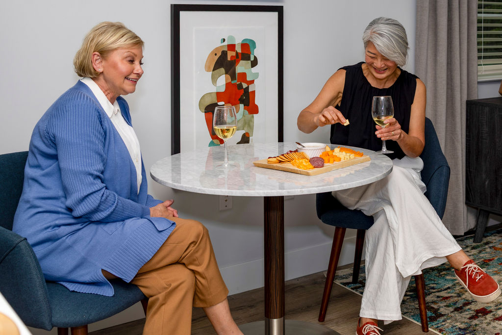 Two women sit at a small round table with wine and a cheese platter, smiling and chatting in a cozy, modern room at Verena at Hilliard, with abstract art on the wall behind them.