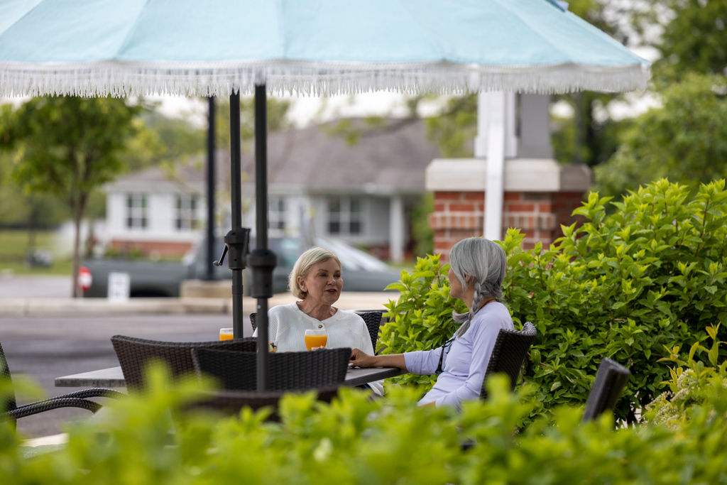 Two older women sit at an outdoor café table under a large blue umbrella at Verena at Hilliard, enjoying drinks and conversation. Green bushes surround them, with a building and parked cars in the background.