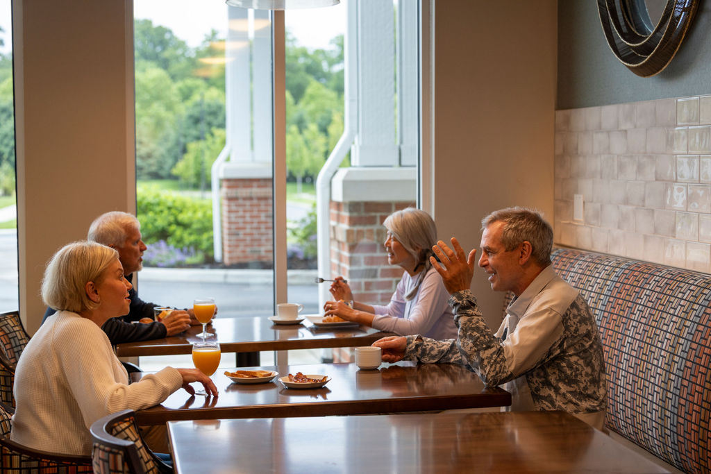 Four older adults sit in pairs at a café table by large windows at Verena at Hilliard, chatting and enjoying drinks and pastries in a cozy, well-lit setting. Trees and a building are visible outside.