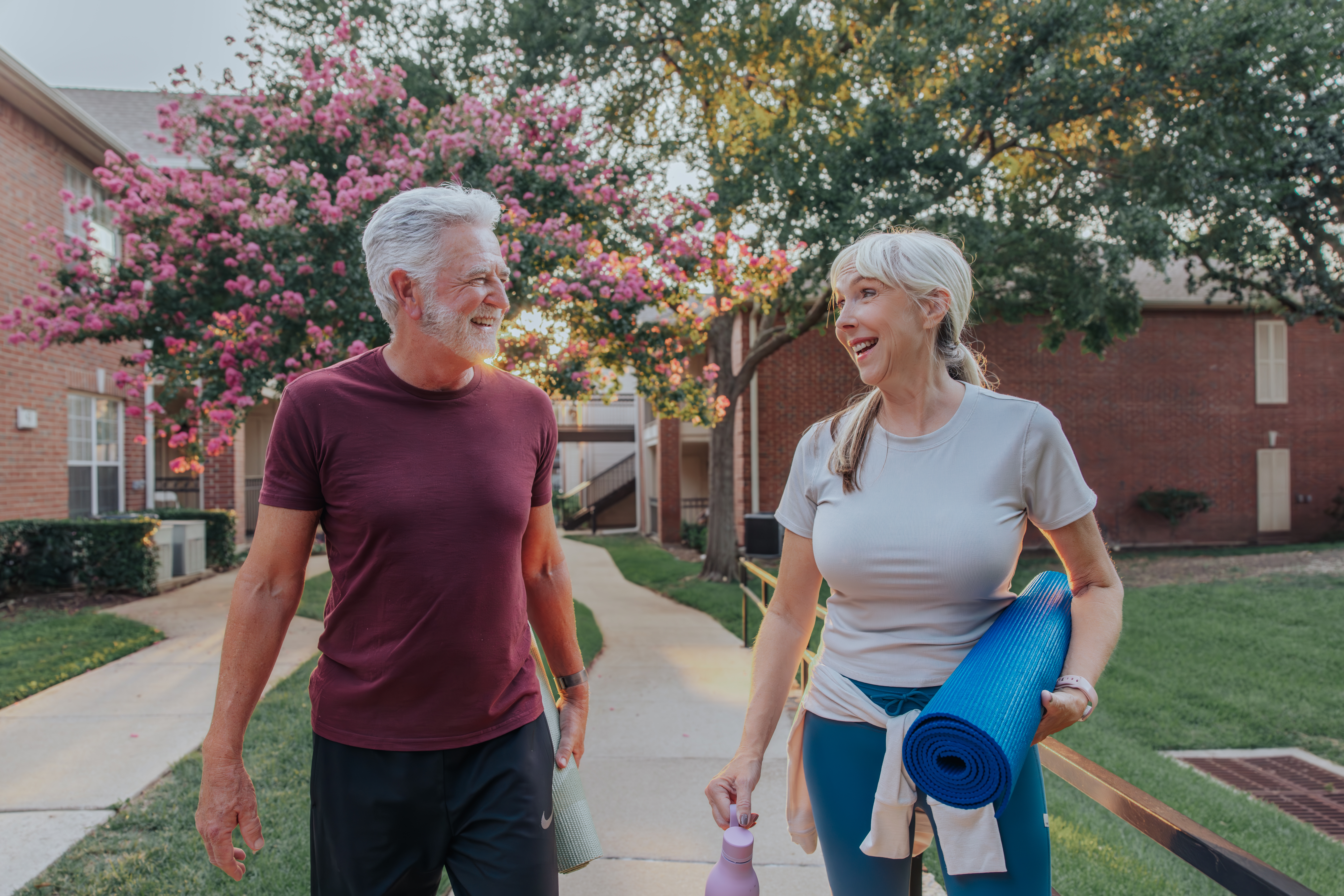 An older man and woman walk outside on a sidewalk at Verena at Hilliard, smiling at each other. The woman holds a rolled yoga mat and water bottle. Dressed in activewear, they stroll past flowers and trees in the background.
