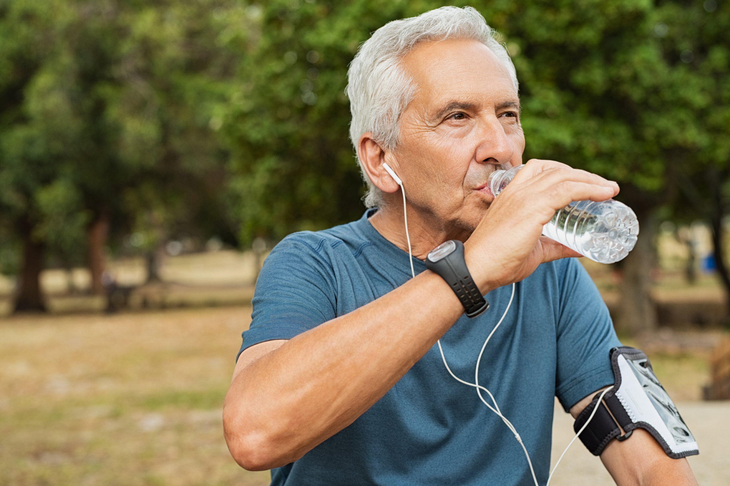 Older man with gray hair wearing a blue shirt, earphones, and a fitness tracker drinks water from a plastic bottle outdoors at Verena at Hilliard, with a smartphone armband on his left arm in the park.