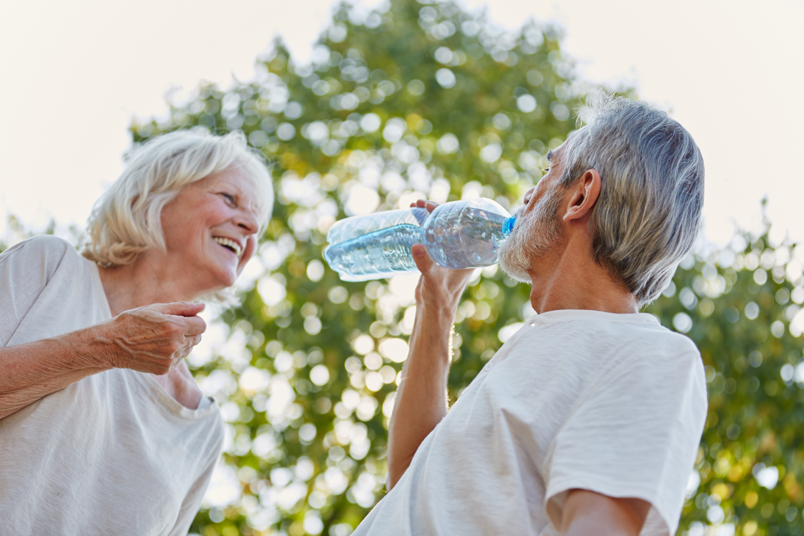 Two older adults outdoors at Verena at Hilliard, both in light-colored shirts. The man drinks from a water bottle while the woman smiles at him, surrounded by sunlit trees in the background on a pleasant, sunny day.