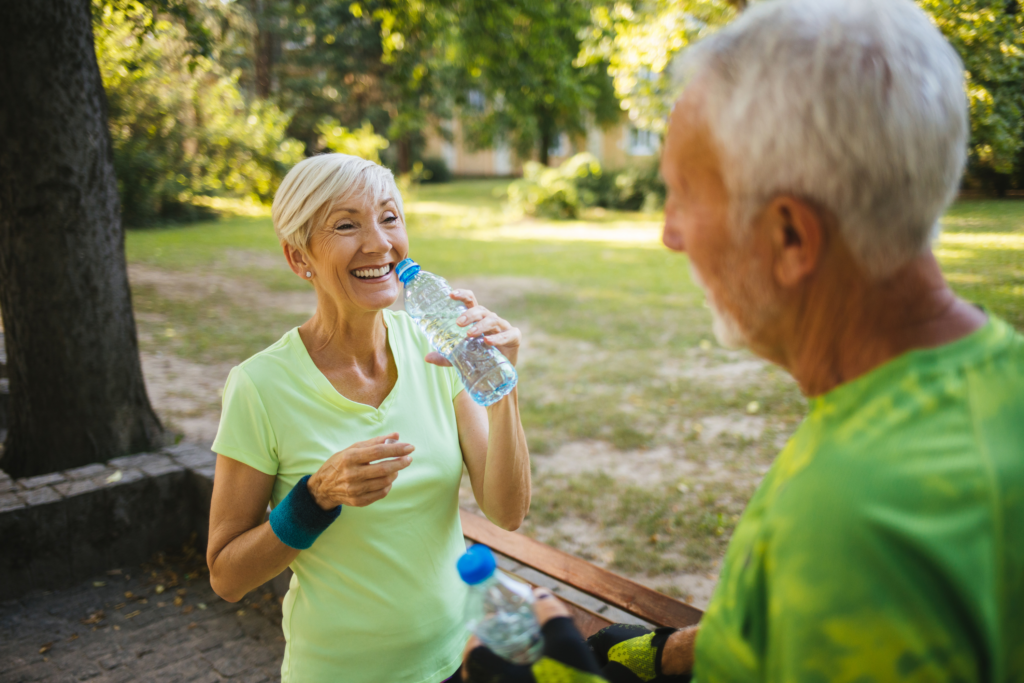 An older woman in a neon green shirt smiles while holding a water bottle in a park at Verena at Hilliard, standing next to an older man also enjoying a break from exercising. Both appear happy and refreshed.
