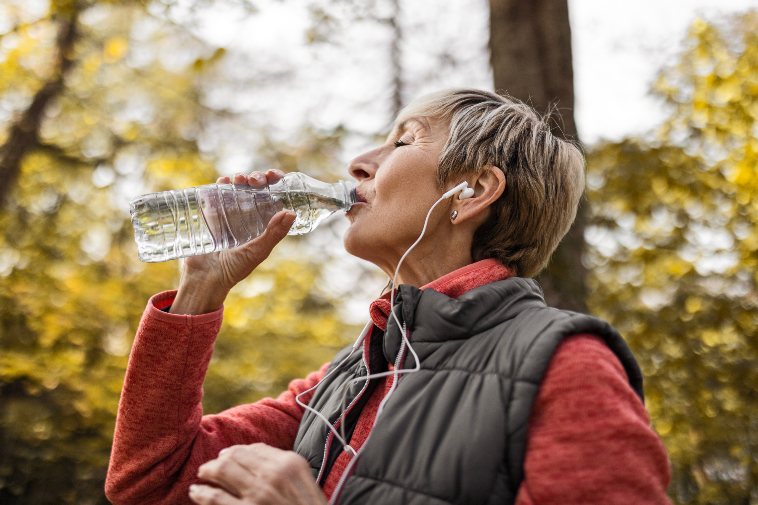 An older woman wearing a red sweater, gray vest, and earphones drinks water from a plastic bottle outdoors at Verena at Hilliard, with trees and golden leaves in the background.