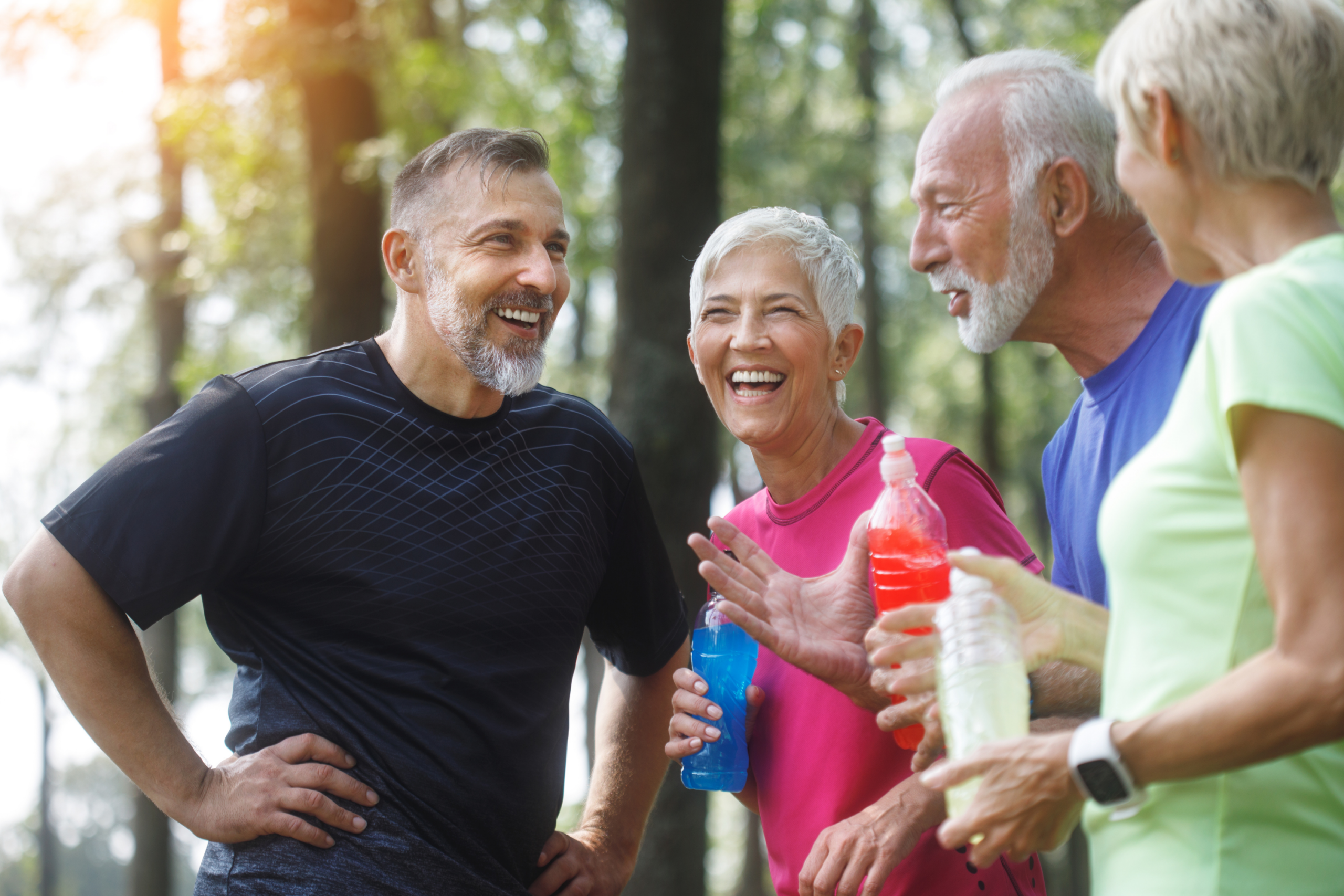 Four older adults in athletic clothing stand outdoors, smiling and laughing while holding water bottles at Verena at Hilliard, with trees and sunlight in the background. They appear to be enjoying a break from exercise.